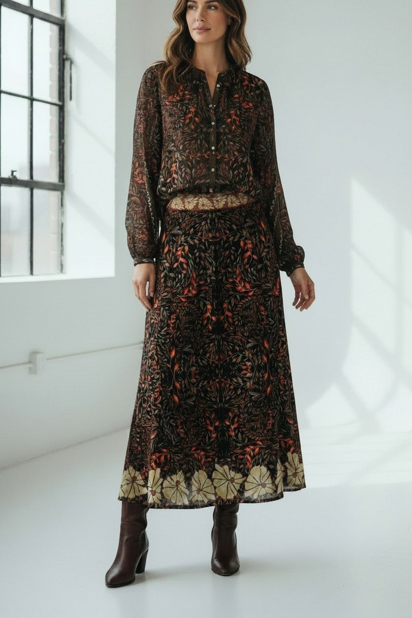 A woman stands in a bright room with large windows and white walls, dressed in a long-sleeved, patterned dress in autumn hues paired with brown heeled boots. Her look is elevated by the addition of the FRANCO FERRARI JEMMA WOOL SKIRT, featuring an elegant botanical print that infuses her ensemble with nature-inspired charm.