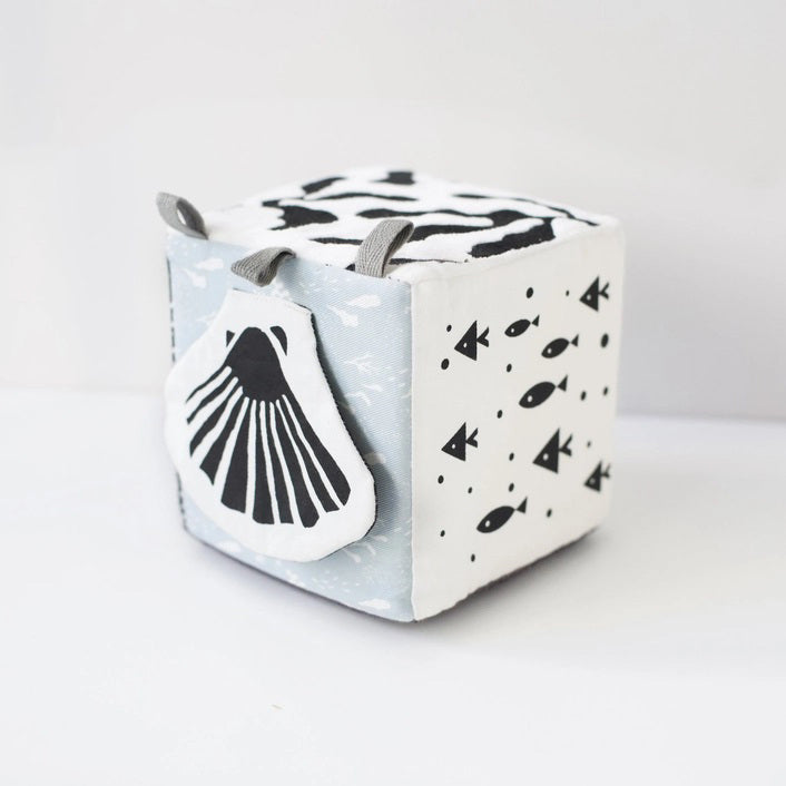 The WEE GALLERY - OCEAN SOFT BLOCK is a monochrome plush cube baby toy featuring high-contrast black and white patterns. Each side of the cube showcases illustrations of ocean life, including a whale and an octopus. Made from soft organic cotton, it also has fabric loops on one corner, making it ideal for infants and toddlers.