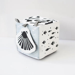 The WEE GALLERY - OCEAN SOFT BLOCK is a monochrome plush cube baby toy featuring high-contrast black and white patterns. Each side of the cube showcases illustrations of ocean life, including a whale and an octopus. Made from soft organic cotton, it also has fabric loops on one corner, making it ideal for infants and toddlers.