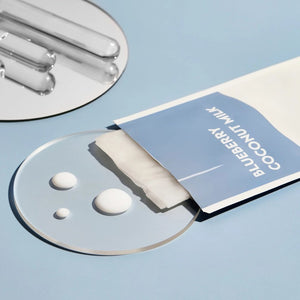 A carton labeled "ESW BEAUTY - BLUEBERRY COCONUT MILK MASK" by ESW BEAUTY sits among a milk bottle, blueberries, coconuts, and coconut flakes on white—perfect inspiration for a blueberry coconut plant-based sheet mask.