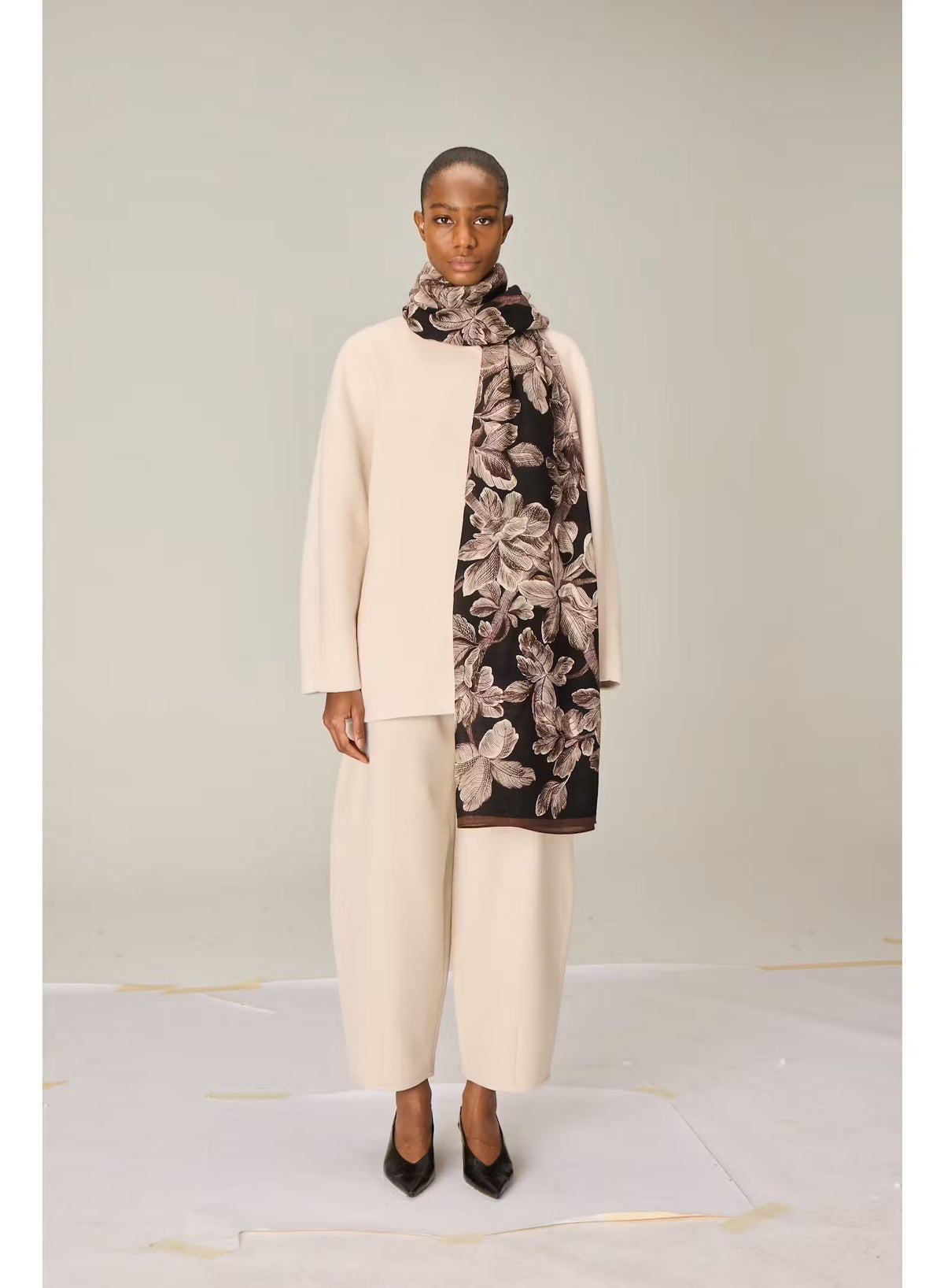 A person stands facing forward, dressed in a cream-colored, loose-fitting outfit featuring wide-legged pants and a long-sleeved top. They have paired their ensemble with black pointed shoes and the INOUI EDITIONS - ARBORIS WOOL SCARF IN BLACK by INOUI, which drapes elegantly over one arm. Made in India from 100% wool, the scarf adds a touch of luxurious comfort against the plain light background.