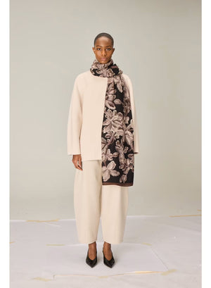 A person stands facing forward, dressed in a cream-colored, loose-fitting outfit featuring wide-legged pants and a long-sleeved top. They have paired their ensemble with black pointed shoes and the INOUI EDITIONS - ARBORIS WOOL SCARF IN BLACK by INOUI, which drapes elegantly over one arm. Made in India from 100% wool, the scarf adds a touch of luxurious comfort against the plain light background.