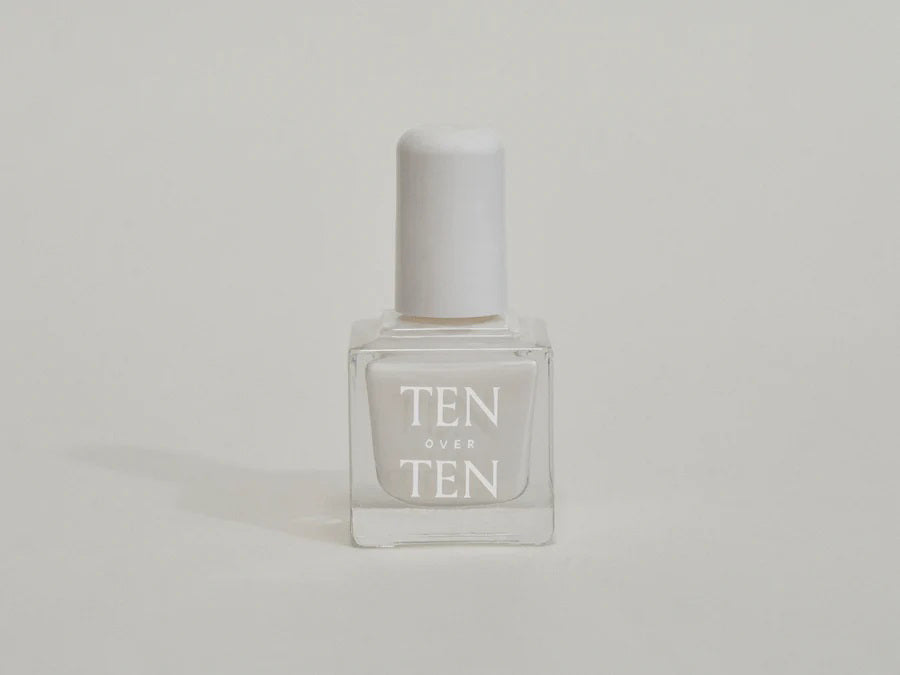 A glass bottle of TENOVERTEN - NAIL POLISH from the brand TEN OVER TEN, featuring a white cap and a dusty pink nail color. The front label displays "TEN OVER TEN" in white uppercase letters. This vegan, eight-free formula ensures both beauty and peace of mind. The background is plain and light-colored.