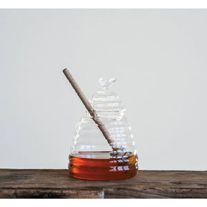 The CREATIVE COOP GLASS BEES NEST HONEY JAR is a beehive-shaped clear glass honey pot, featuring a lid topped with a small glass bee and comes with a wood honey dipper inside.