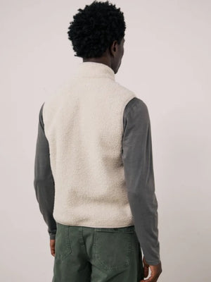 A man is wearing the HARTFORD - BASTA SHERPA VEST IN NATURAL over a gray long-sleeve shirt, holding the vest’s zipper with one hand. The plain, light-colored background complements the cozy, Hartford-inspired style of this men’s vest from HARTFORD.