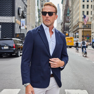 A man wearing sunglasses, the COLLARS AND CO MAVERICK KNIT UNSTRUCTURED BLAZER IN NAVY over a white shirt, light pants, and brown loafers stands on a sidewalk in front of a light stone building.