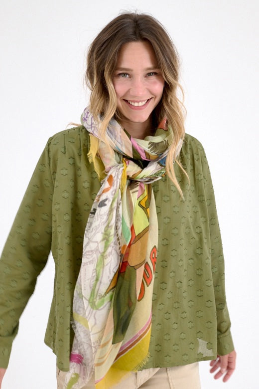 A woman in a green dress drapes the STORIATIPIC Gabie Scarf in Rose over her shoulders. This luxury silk accessory by STORIATIPIC features playful illustrations, maps, and "TOUR DE FRANCE" on a light background.