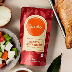 A packet of BRODO BROTH COMPANY’s BRODO BROTH- SIGNATURE HEARTH BONE BROTH sits on a light surface, surrounded by a bowl of chopped vegetables, a cooked turkey leg, half an onion, a bay leaf, and a cup of rich bone broth made from grass-fed beef and organic chicken.