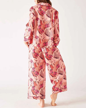 A woman stands barefoot wearing the MERSEA Over The Moon PJ Set in Rosewood by MERSEA—light pink cotton pajamas with vibrant pink and purple florals, a button-up shirt, wide-leg pants, and a comfy stretch waistband. Her face is partially out of frame.