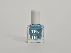 A glass bottle of TENOVERTEN - NAIL POLISH from the brand TEN OVER TEN, featuring a white cap and a dusty pink nail color. The front label displays "TEN OVER TEN" in white uppercase letters. This vegan, eight-free formula ensures both beauty and peace of mind. The background is plain and light-colored.