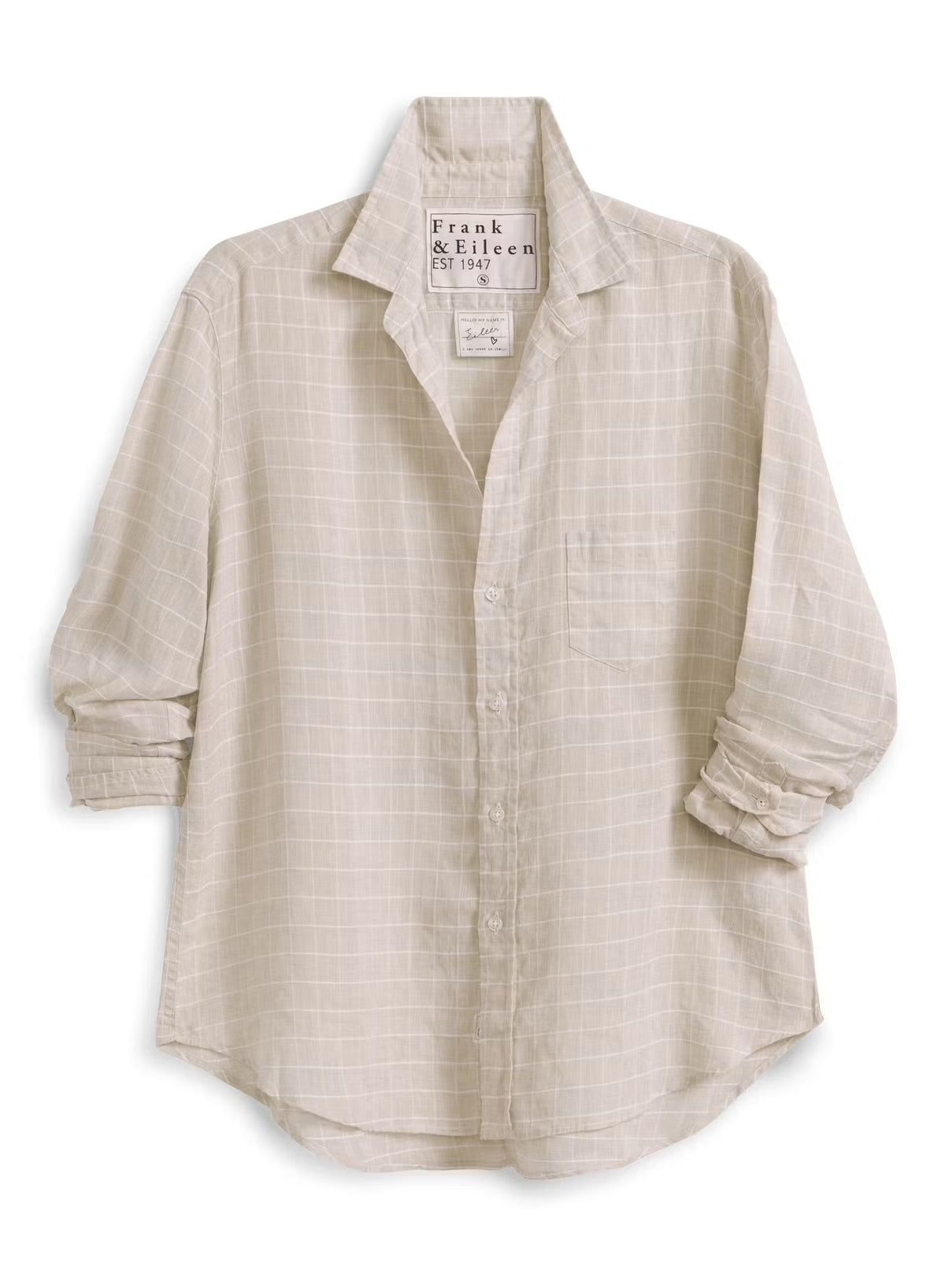 A woman with brown hair, smiling on a sunlit cobblestone street with blurred outdoor café tables in the background, wears the FRANK & EILEEN EILEEN Relaxed Button Up in Italian Cotton Tan with Textured Grid paired with jeans.