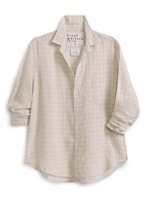 A woman with brown hair, smiling on a sunlit cobblestone street with blurred outdoor café tables in the background, wears the FRANK & EILEEN EILEEN Relaxed Button Up in Italian Cotton Tan with Textured Grid paired with jeans.