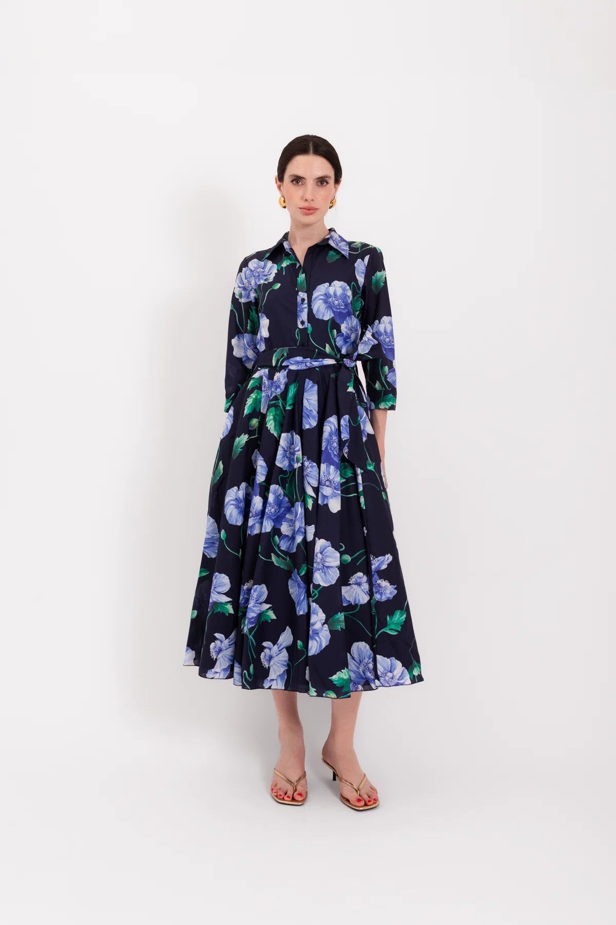A woman stands with her back to the camera, wearing the elegant LAVI - SPRITZ BLUE POPPIES DRESS by LAVI, featuring a stunning blue midi silhouette decorated with purple and blue poppy flowers. She has short dark hair, gold earrings, and open-toed heels against a plain white background. This striking piece is beautifully made in Italy.