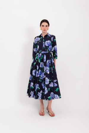 A woman stands with her back to the camera, wearing the elegant LAVI - SPRITZ BLUE POPPIES DRESS by LAVI, featuring a stunning blue midi silhouette decorated with purple and blue poppy flowers. She has short dark hair, gold earrings, and open-toed heels against a plain white background. This striking piece is beautifully made in Italy.