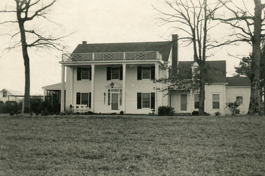 Black-and-white photo of a large, two-story Colonial-style house with white siding, a columned porch, shutters, and a balcony. Leafless trees surround the house, and a grassy lawn stretches in front.