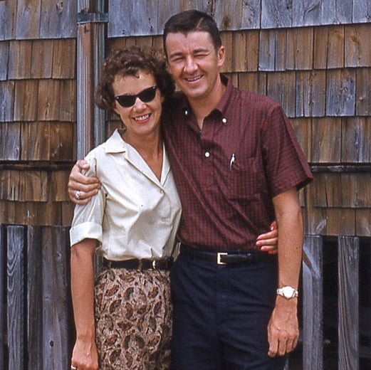 A smiling woman in sunglasses and a patterned skirt stands with her arm around a smiling man in a short-sleeve maroon shirt. They are posing together in front of a wooden shingle wall.
