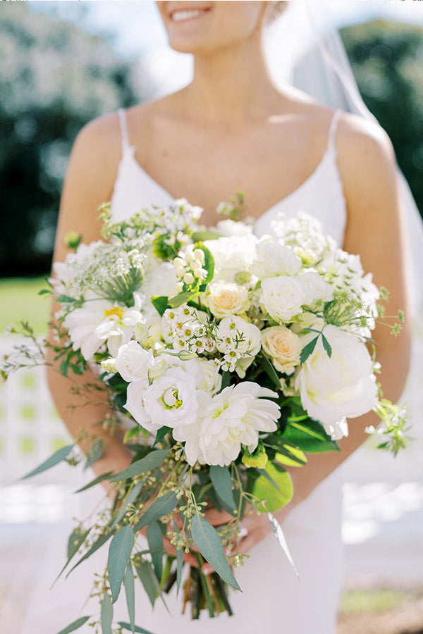 A bride in a white dress holds a large bouquet of white and cream flowers, including roses and greenery, outdoors on a sunny day.