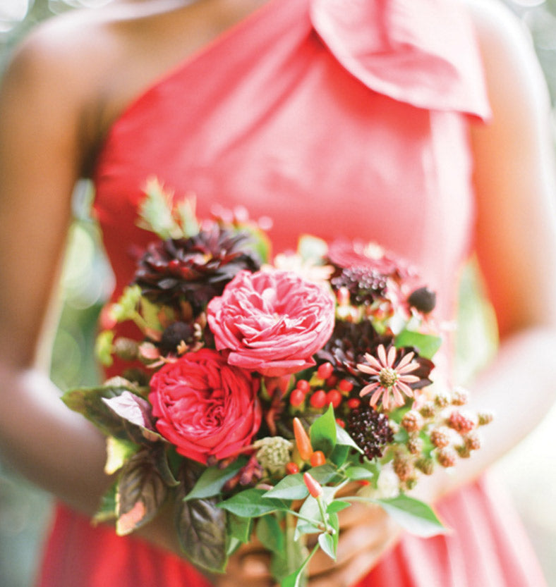 A person in a pink one-shoulder dress holds a vibrant bouquet of red, pink, and dark burgundy flowers, accented with green leaves and berries, against a softly blurred outdoor background.
