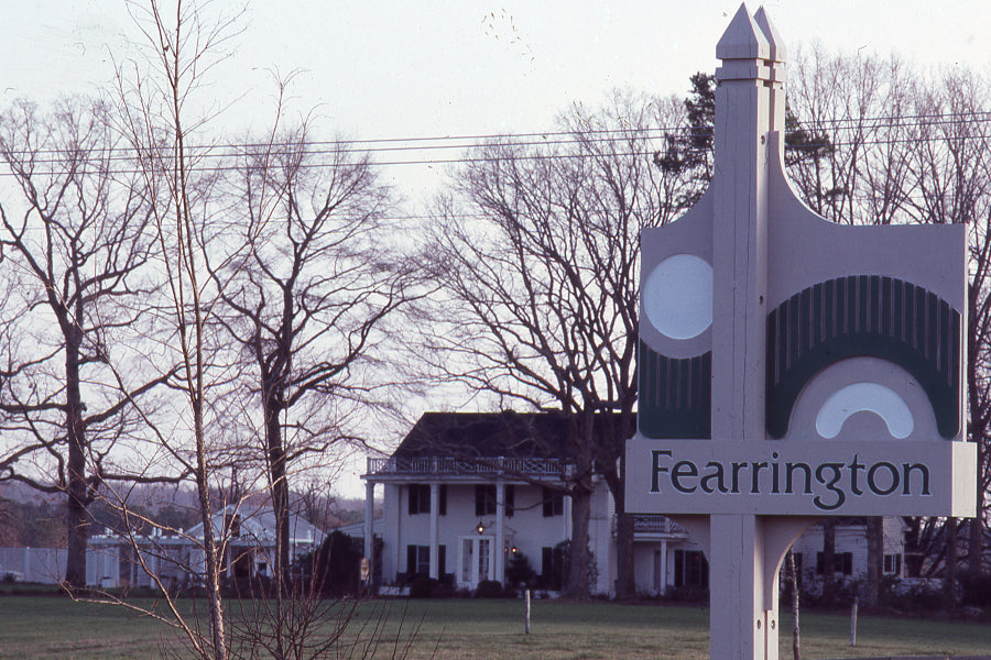 A large “Fearrington” sign stands in front of leafless trees and a white, two-story house with columns, on a grassy area under a pale sky.