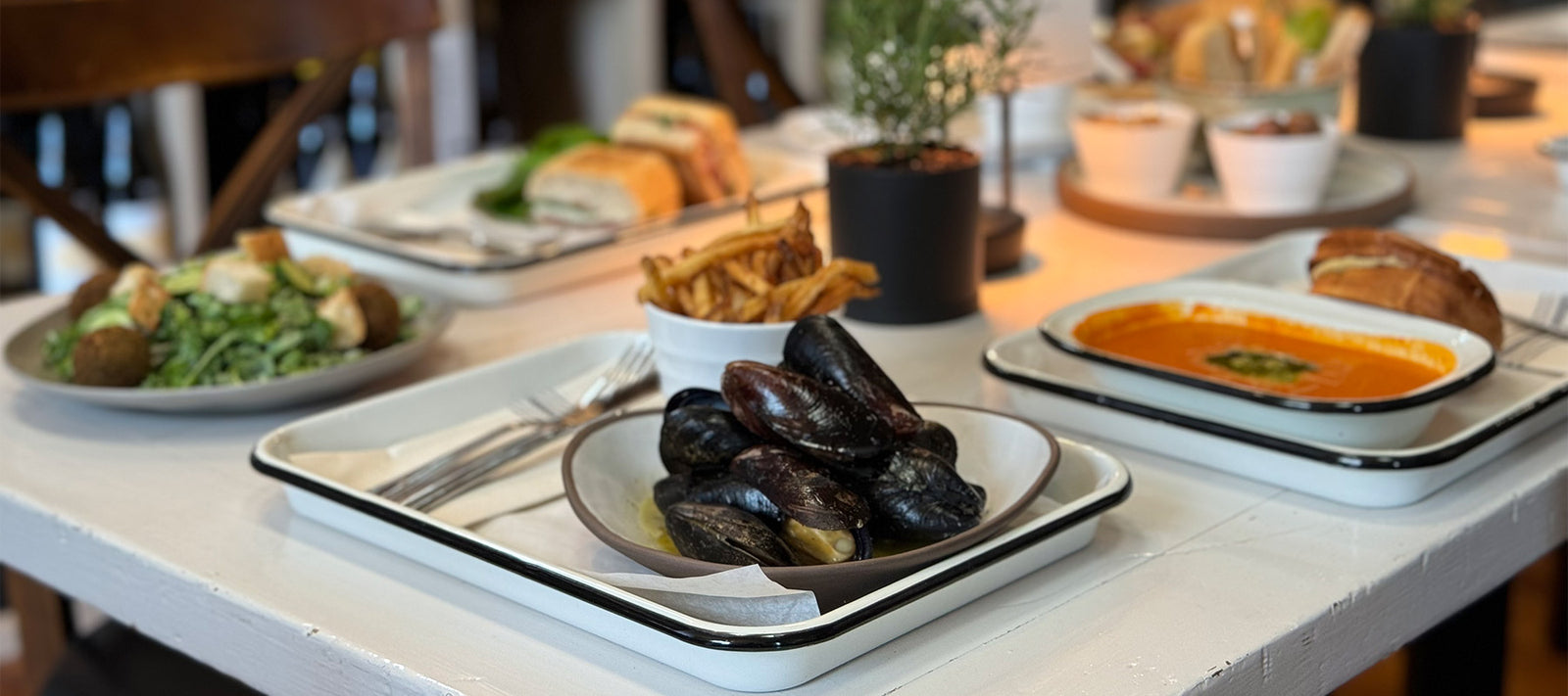 A close-up of a table set with dishes including a bowl of mussels, a bowl of fries, tomato soup with herbs, salad, and sandwiches, with potted plants as centerpieces.