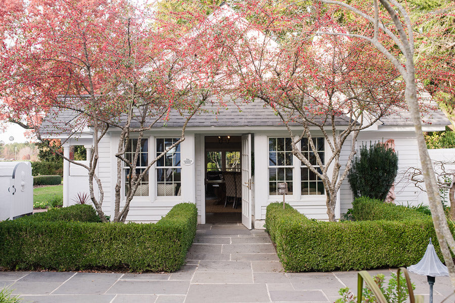 A charming white cottage with large windows and a gray roof is framed by two small trees with red leaves and neatly trimmed hedges along a stone pathway leading to the entrance.