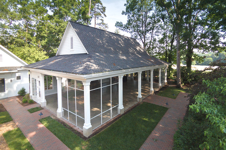 A large, open-sided pavilion with a steeply pitched roof and white columns stands on a brick patio, surrounded by grass and trees. The structure features screened-in sides and is adjacent to a wooded area.
