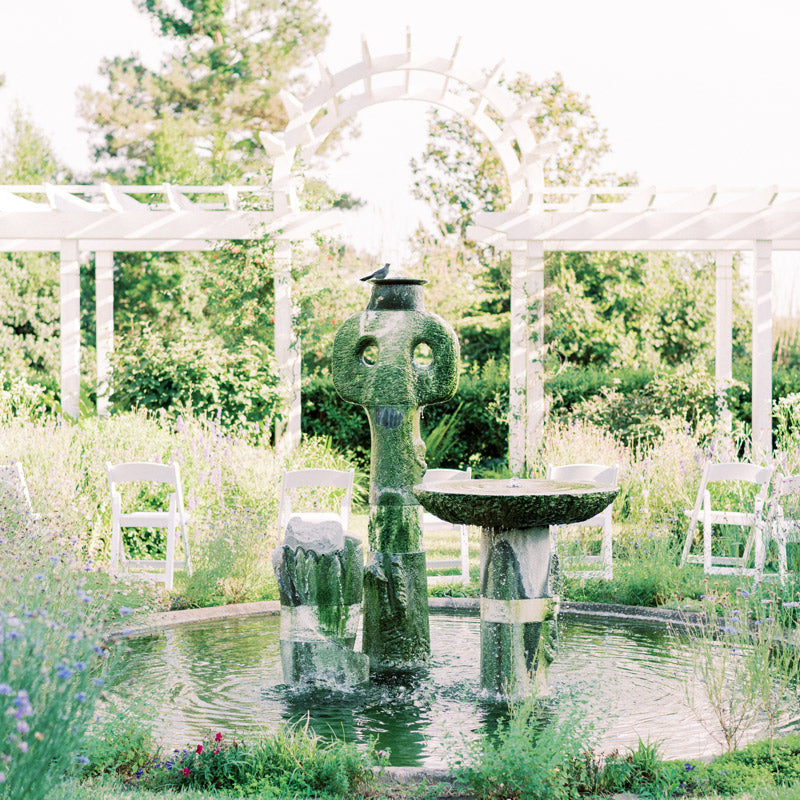 A stone fountain with moss and water features stands in the center of a garden, surrounded by greenery, white pergolas, and white folding chairs under a bright, sunny sky.