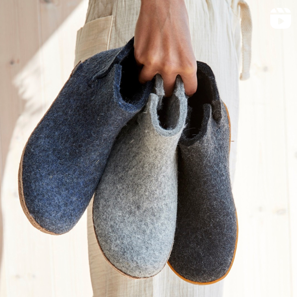 A person wearing cream-colored pants holds three pairs of wool felt slippers in dark blue, light gray, and dark gray, by their heels. The background is light-colored wood paneling.