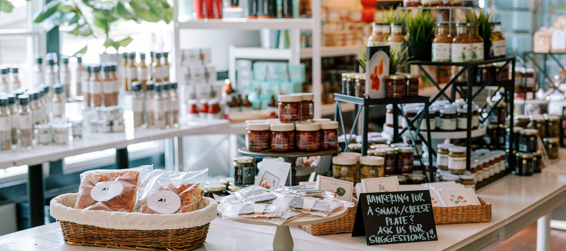 A specialty food store display with shelves of jars, bottles, and packages of gourmet items. A basket of bread and a chalkboard sign offering snack and cheese platter suggestions are on a white counter in the foreground.