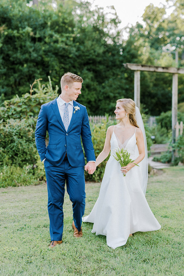 A bride and groom walk hand in hand outdoors on grass, smiling at each other. The bride wears a white dress and holds a bouquet, while the groom wears a blue suit. Greenery and a wooden arbor are visible in the background.