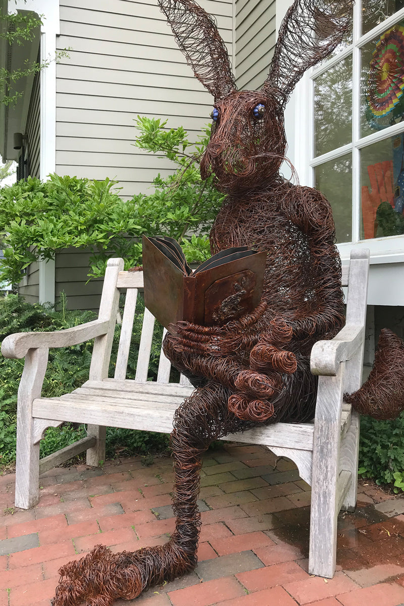 A large wire sculpture of a rabbit sits upright on a wooden bench outside a house, holding an open book as if reading. The sculpture has exaggerated ears and is surrounded by greenery and brick pavement.