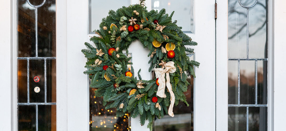 A festive holiday wreath with green pine branches, orange slices, red berries, and a cream ribbon bow hangs on a white door with decorative glass panels.