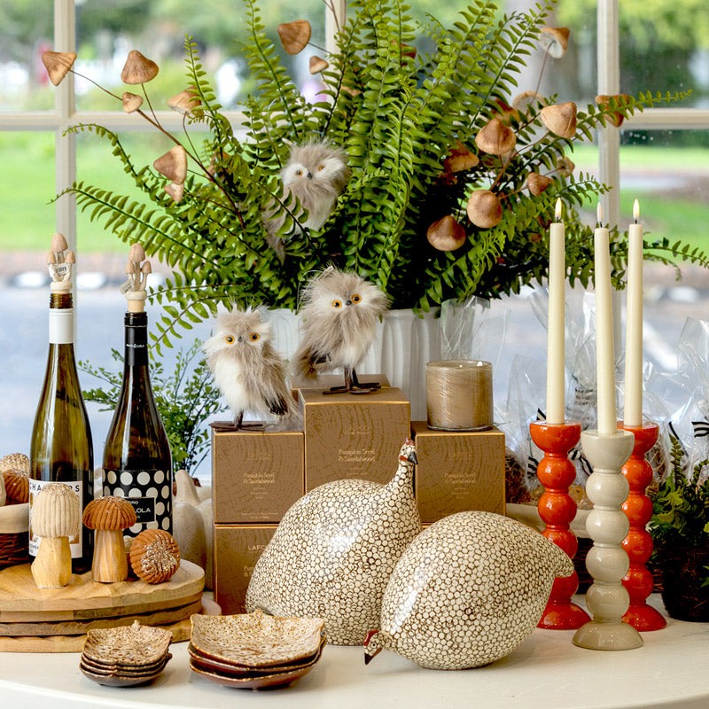 A decorative display features ceramic guinea fowl, owl figurines, ferns, candles, wine bottles, stacked boxes, mushroom ornaments, and small dishes arranged on a table in front of a window.