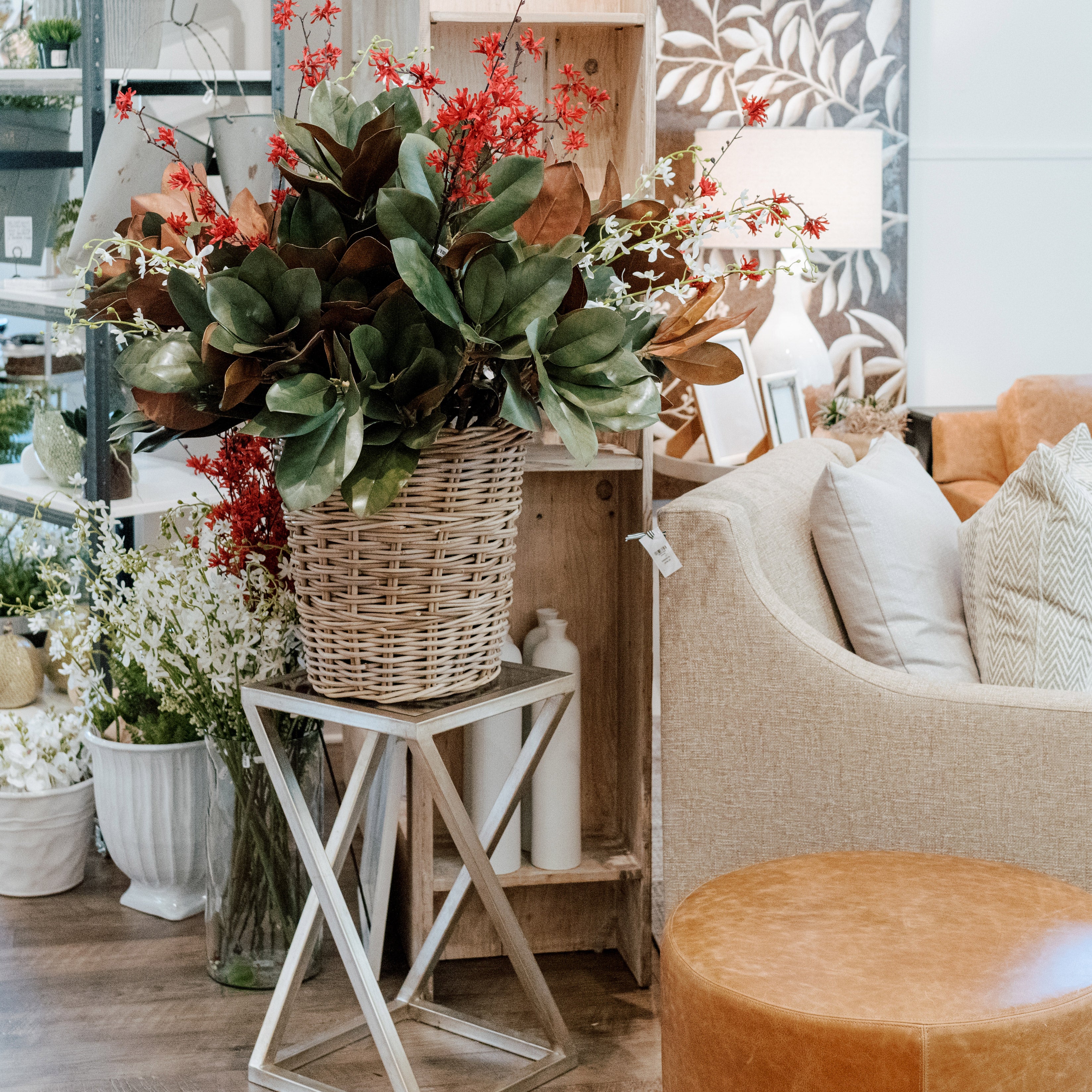 A large wicker basket filled with green leaves and red flowers sits on a modern stand in a cozy, stylish living room with a beige sofa, floral decor, and neutral furnishings.