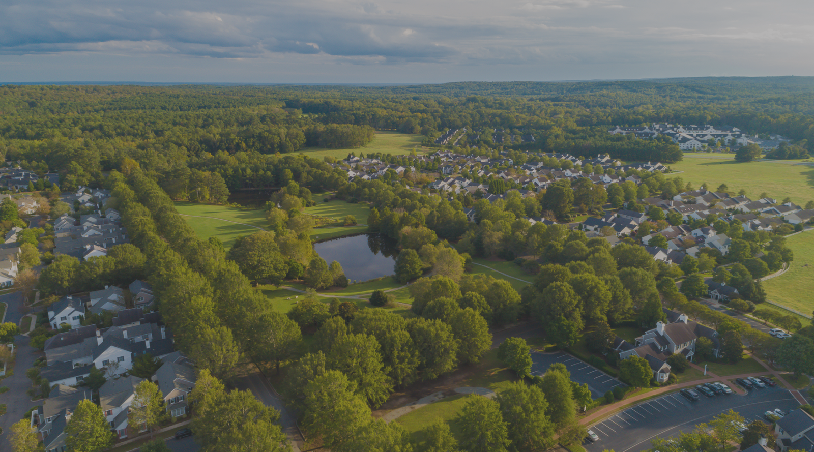 Aerial view of a suburban neighborhood surrounded by dense green trees, a pond, open grassy fields, and clusters of houses under a partly cloudy sky.