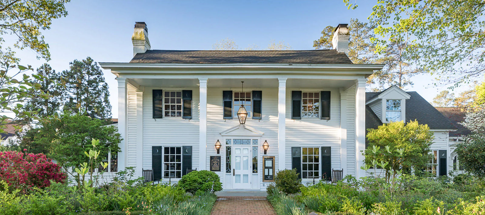A white, two-story colonial-style house with black shutters, columns, and a centered front door, surrounded by green shrubs, flowers, and trees under a clear blue sky.