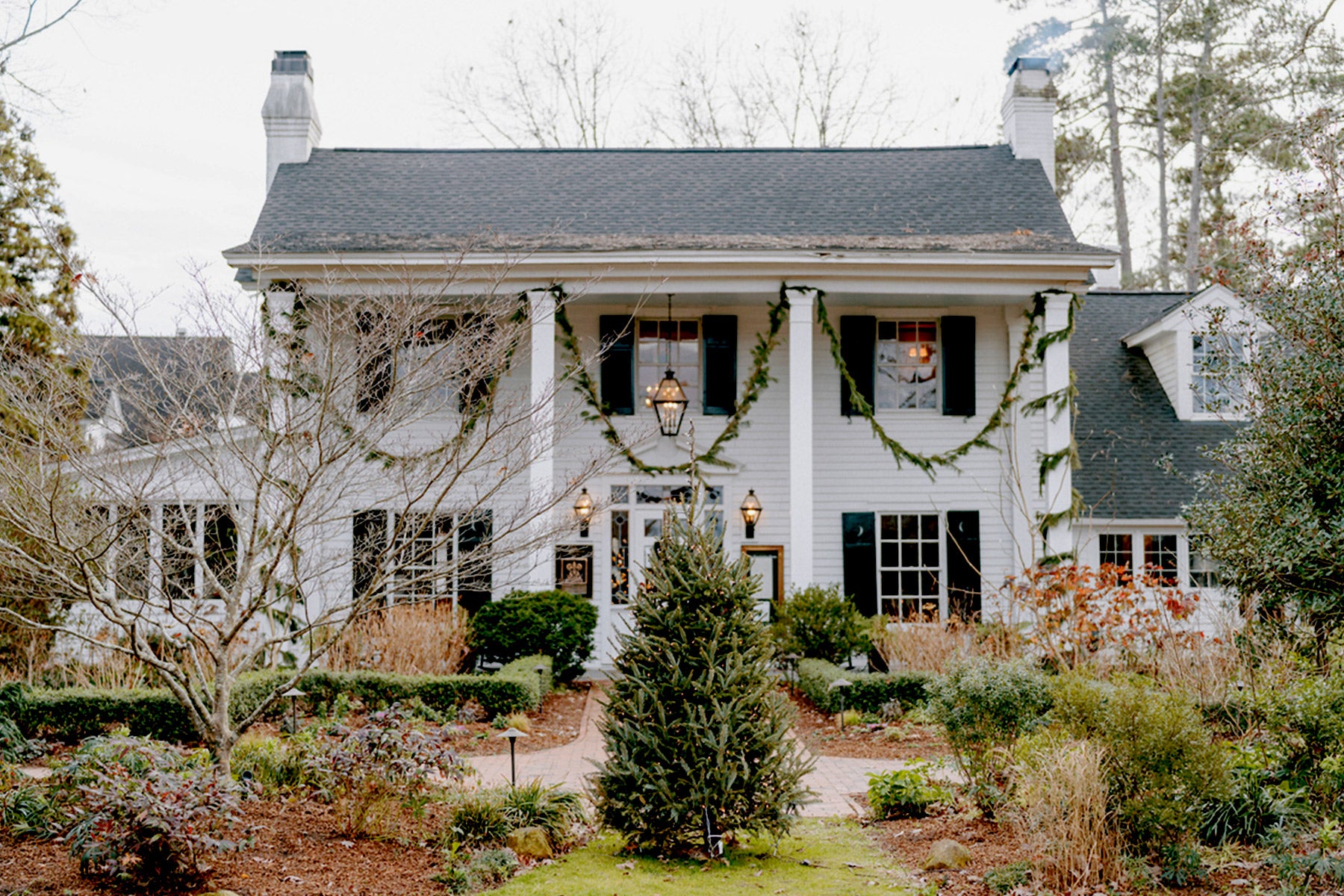 A large white two-story house with black shutters, decorated with green garland. Leafless trees and plants surround a walkway leading to the front door. The yard contains winter foliage and a small evergreen tree.