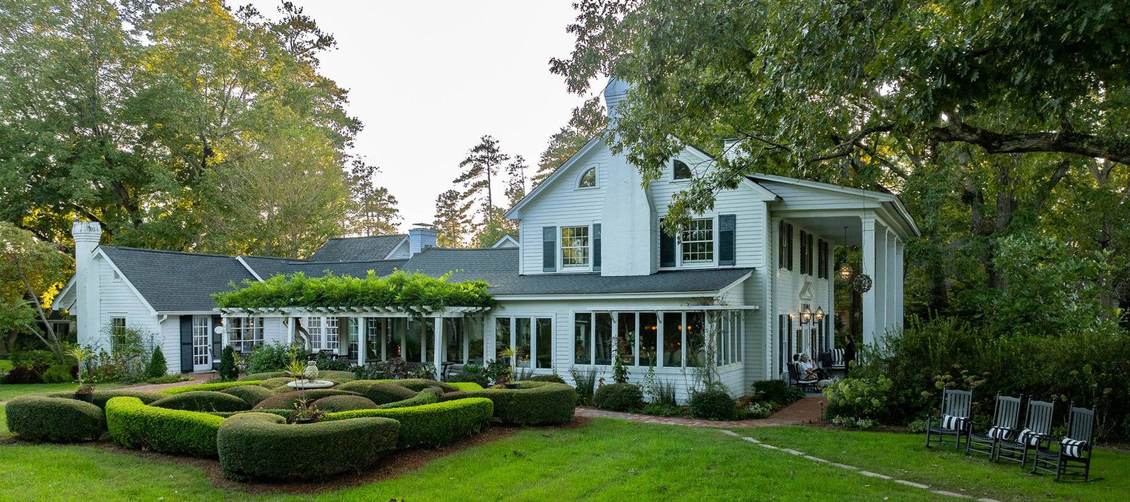 A large white house with black shutters and a covered porch sits amid lush greenery and manicured bushes, with rocking chairs on the side porch and a circular hedge garden in front. Tall trees surround the property.