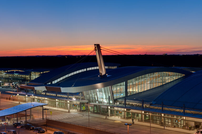 A modern airport terminal with curved roofs is shown at dusk. The sky glows with shades of orange and blue as the sun sets behind the building, and soft lights illuminate the terminal's glass facade.