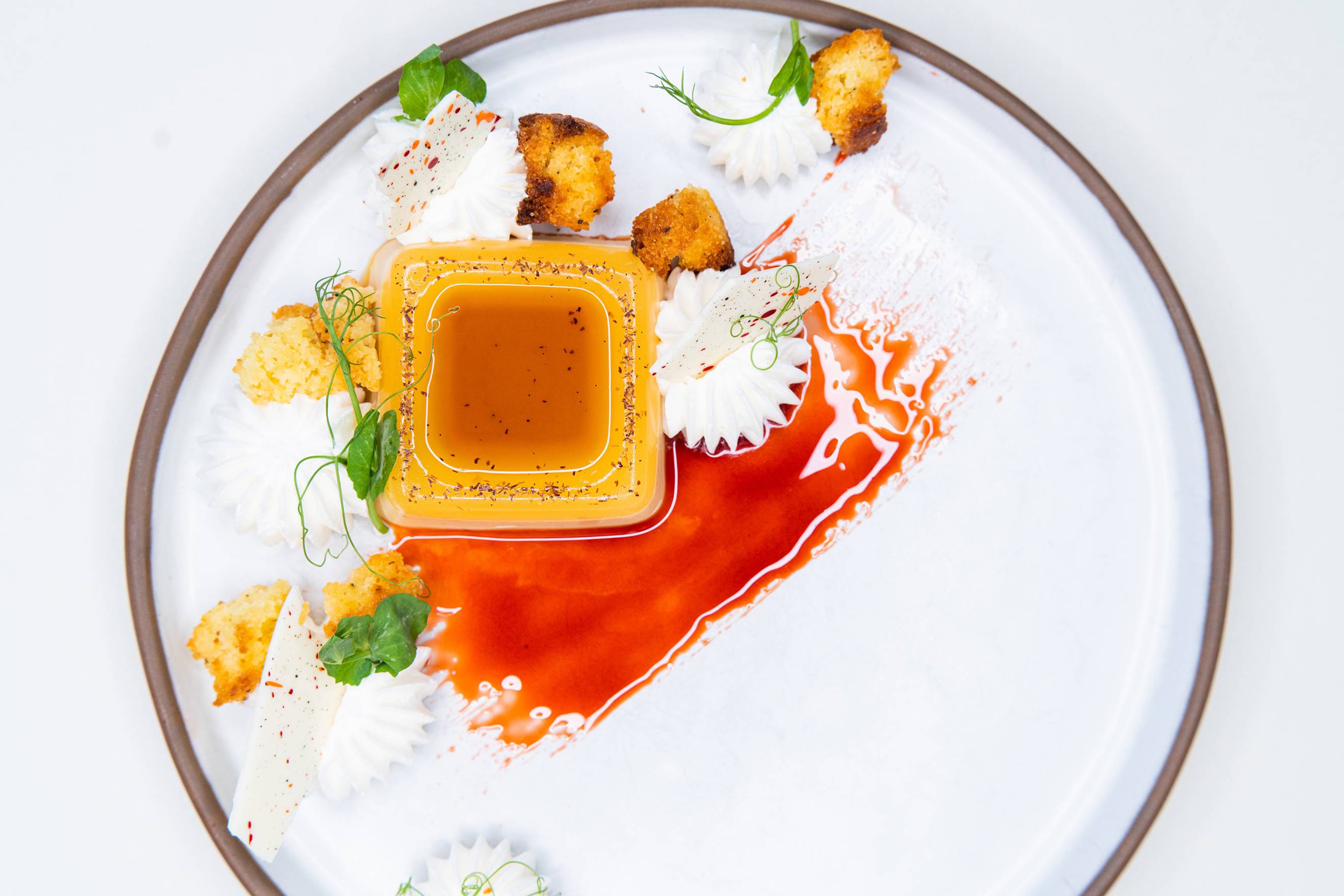 A gourmet dessert with a square flan topped with caramel sauce, surrounded by whipped cream swirls, croutons, green herbs, and a smear of red sauce on a white plate.