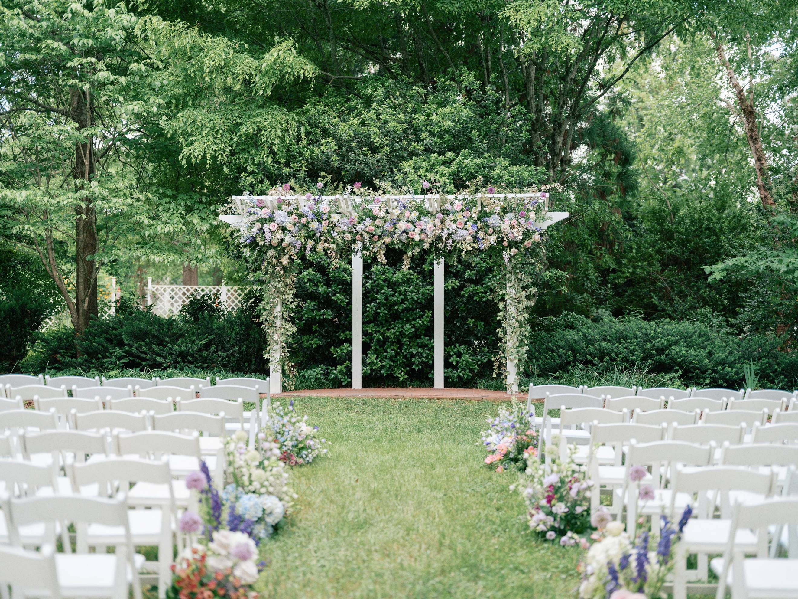 An outdoor wedding ceremony setup with rows of white chairs facing a white pergola decorated with lush, pastel flowers, surrounded by greenery and trees. Floral arrangements line the aisle on a grassy lawn.