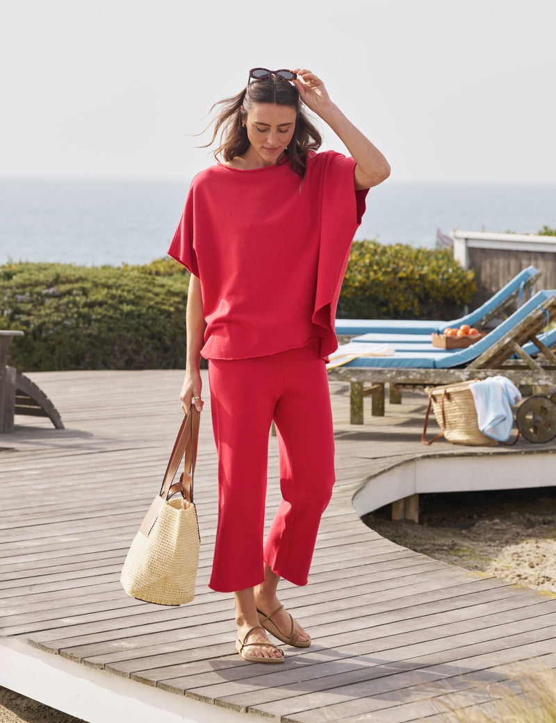 A woman wearing a loose red top and matching red pants walks on a wooden deck near the ocean. She carries a straw tote bag, wears sandals, and adjusts her sunglasses. Lounge chairs and greenery are in the background.