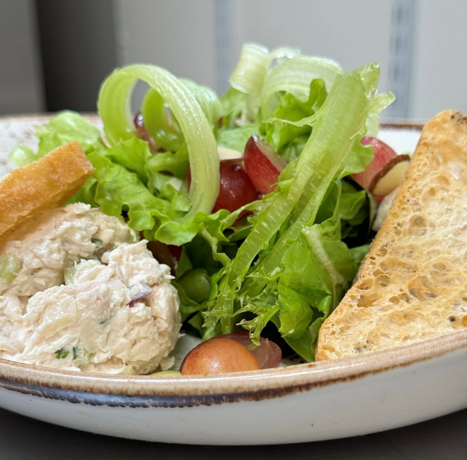 A plate with chicken salad, lettuce, sliced celery, red grapes, almond slivers, and two pieces of toasted bread. The dish is fresh and colorful, served in a rustic bowl.