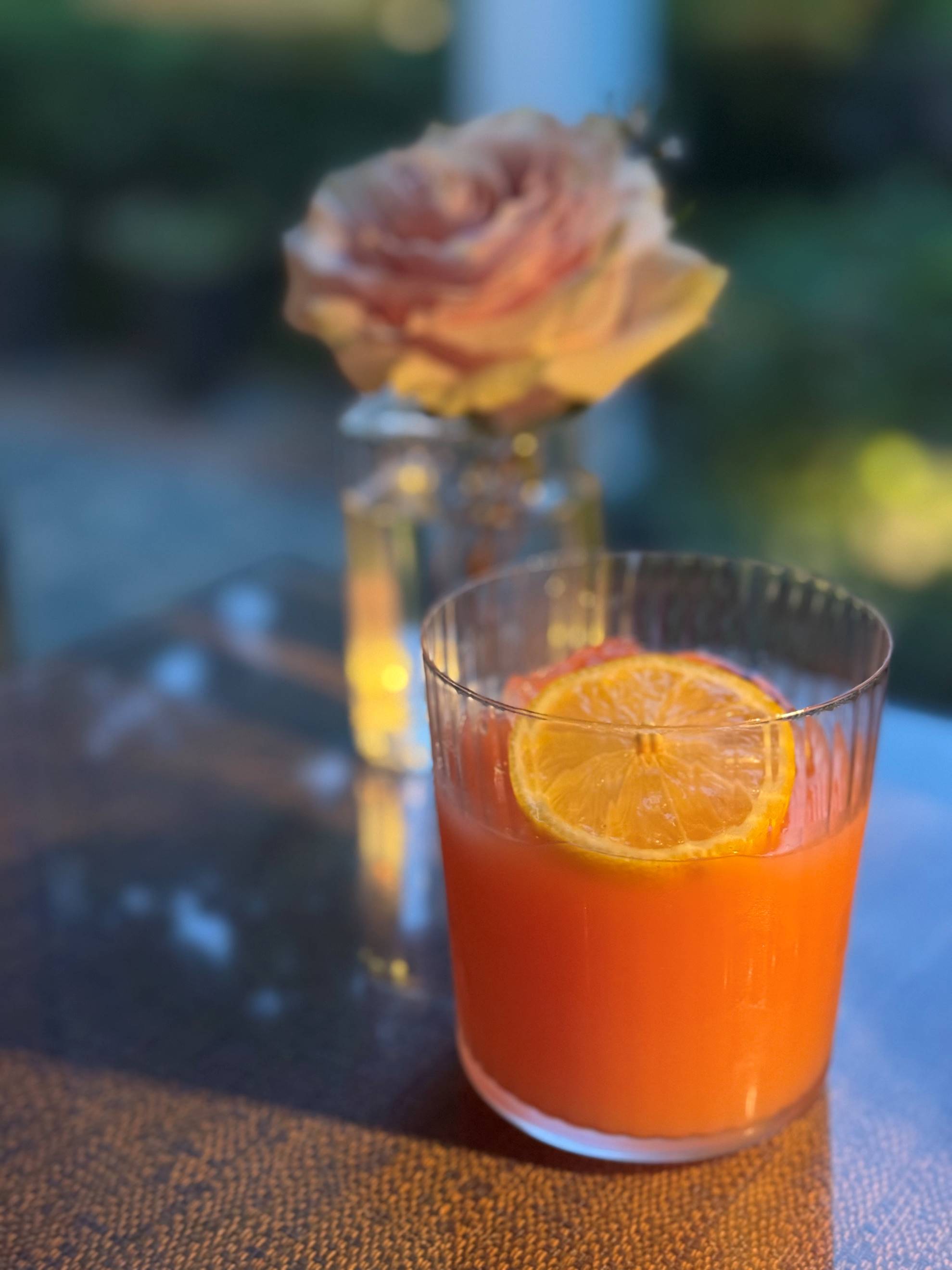 A glass of orange juice with a lemon slice sits on a table, with a blurred pink rose in a small glass vase in the background. Soft sunlight creates a warm, inviting atmosphere.