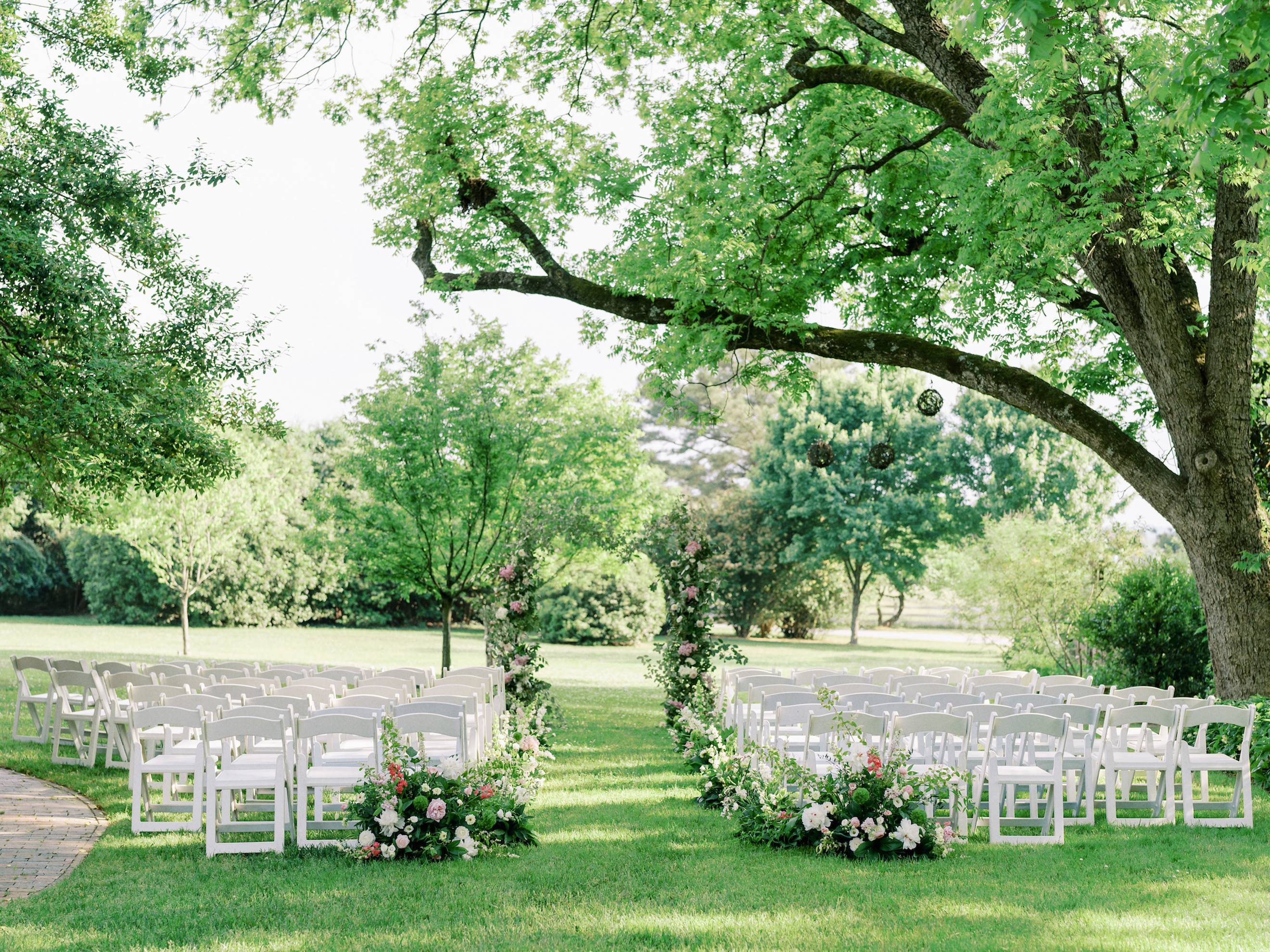 Outdoor wedding ceremony setup with rows of white chairs arranged on a green lawn, decorated with lush floral arrangements and greenery, under large shade trees on a sunny day.