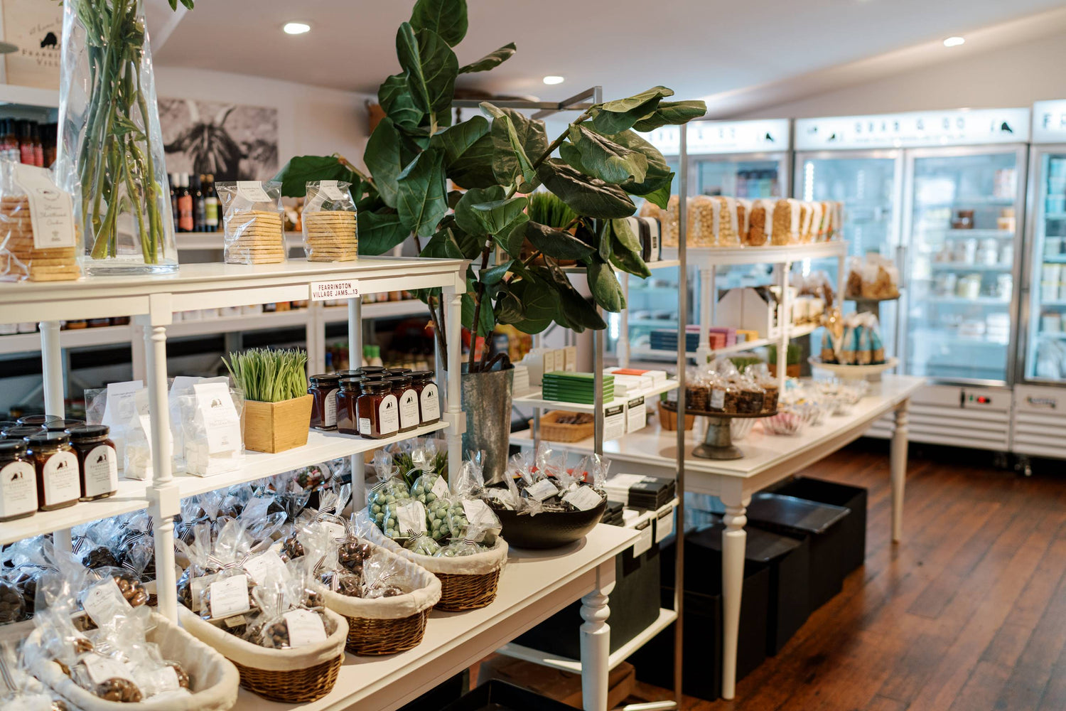 Bright, modern gourmet grocery store with wooden floors, shelves of packaged goods, jars, and baskets of snacks. A large green plant decorates the center. Refrigerated cases line the back wall.