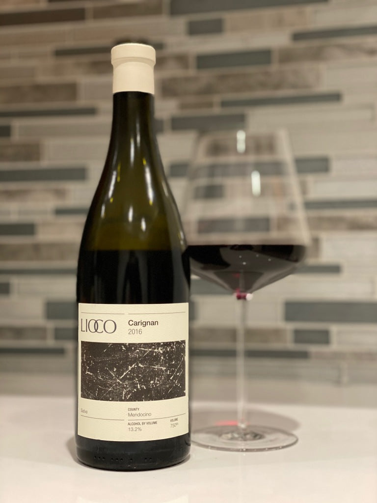 A bottle of Lioco Carignan 2016 wine stands next to a filled wine glass on a white countertop, with a modern, tiled backsplash in the background.