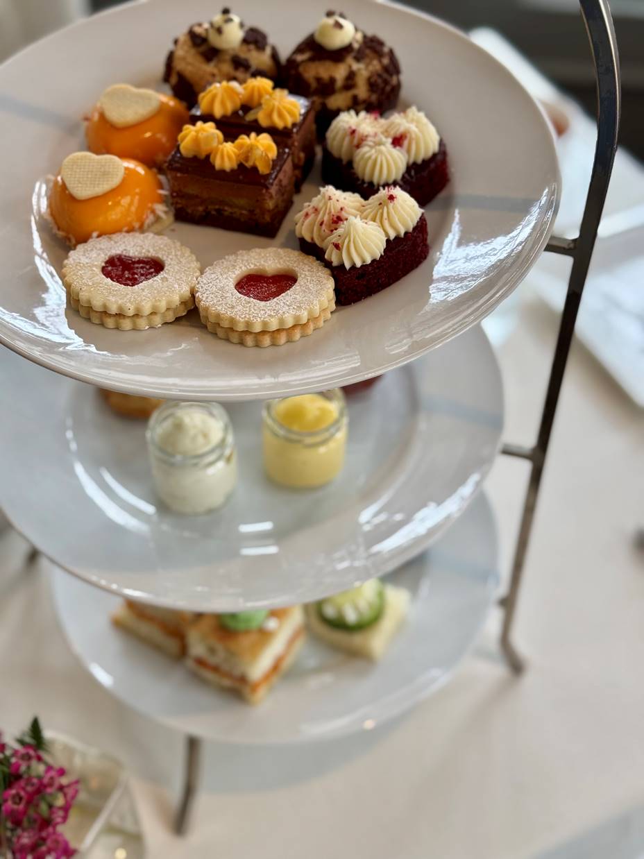 A tiered serving tray holds assorted pastries and cakes on the top plate, jars of cream on the middle plate, and finger sandwiches with cucumber slices on the bottom plate, set on a white tablecloth.