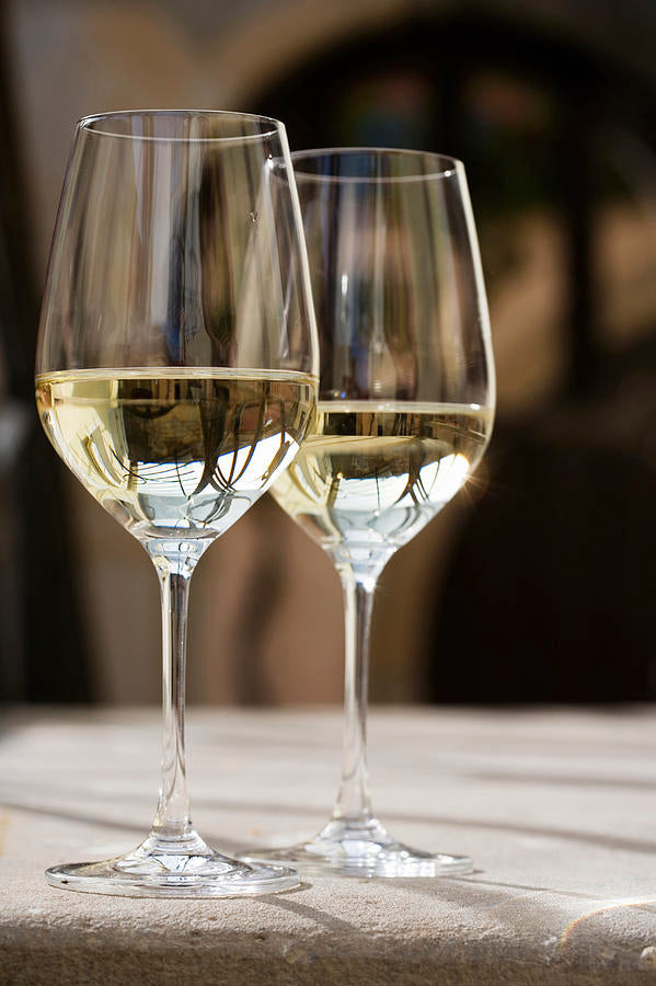 Two clear wine glasses filled with white wine are placed side by side on a light-colored surface, with a soft-focus background suggesting an indoor or outdoor setting.