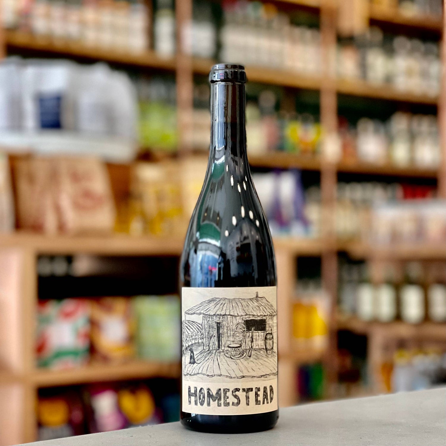 A bottle of wine labeled "Homestead" with a black-and-white drawing of a rustic house, displayed on a counter in front of shelves stocked with various grocery items.